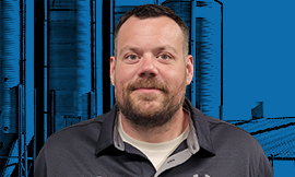 Portrait of a man with short hair and a beard, wearing a dark shirt, standing in front of a blue data-center backdrop.